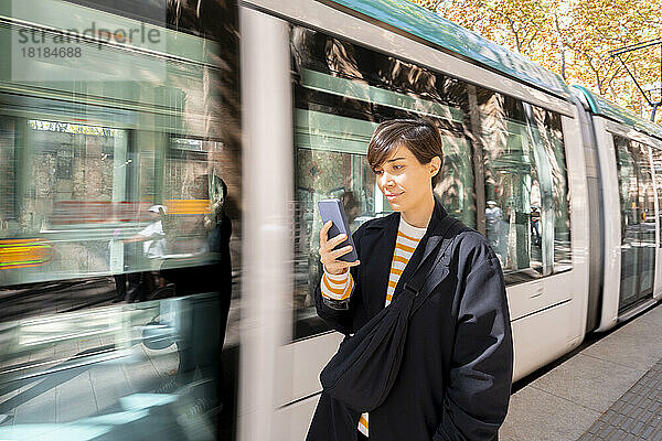 Frau benutzt Smartphone und steht an der vorbeifahrenden Straßenbahn