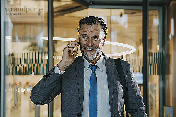 Lächelnder reifer Geschäftsmann  der vor einer Glastür mit dem Smartphone spricht