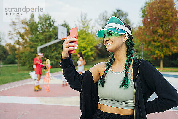 Lächelnde Frau mit Sonnenblende macht Selfie mit dem Handy auf dem Sportplatz