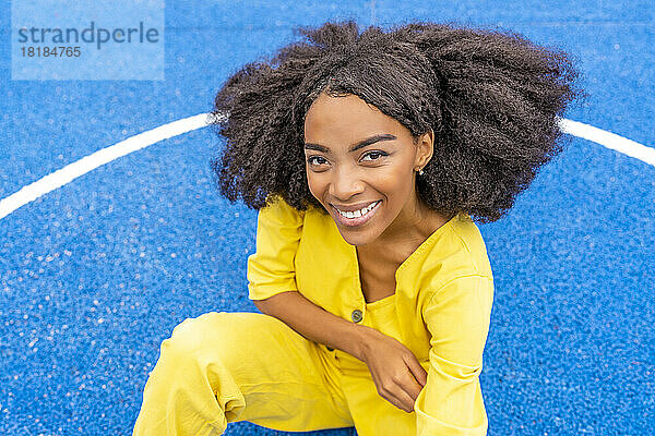 Lächelnde junge Frau sitzt auf dem blauen Basketballplatz