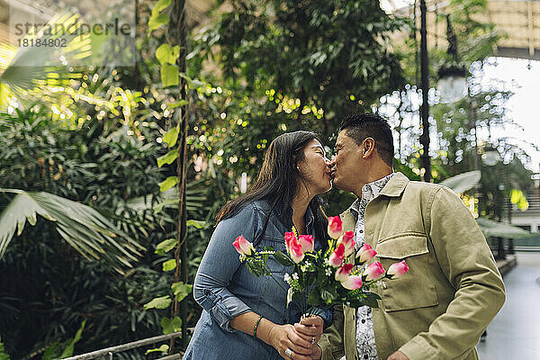 Reife Frau hält Blumen in der Hand und küsst Mann am Valentinstag