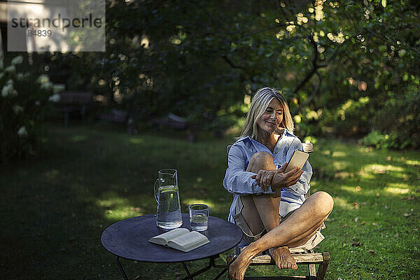Reife Frau sitzt mit Smartphone auf Stuhl im Garten