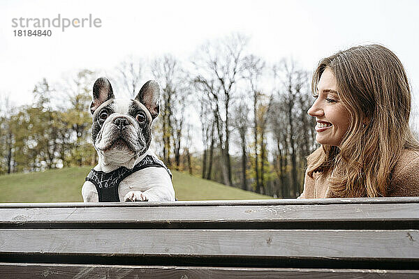 Glückliche Frau mit französischer Bulldogge sitzt auf Bank im Park