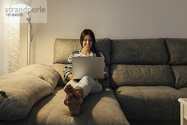 Glückliche Frau benutzt Laptop auf Sofa im Wohnzimmer zu Hause