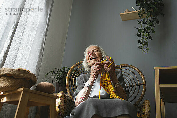 Glückliche Seniorin strickt auf Stuhl im Wohnzimmer
