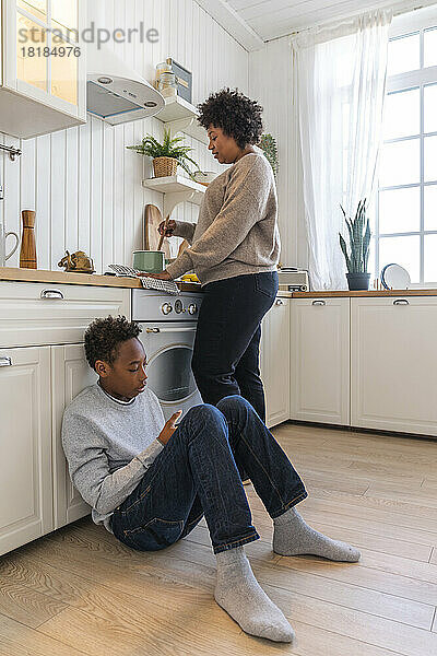 Mutter bereitet Essen zu  während ihr Sohn zu Hause auf dem Boden neben dem Schrank in der Küche sitzt