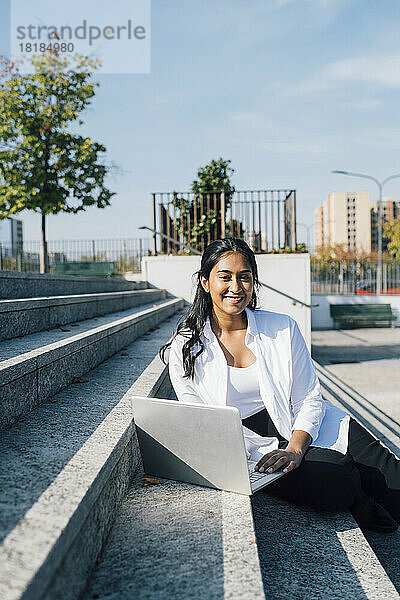 Glückliche junge Frau mit Laptop sitzt an einem sonnigen Tag auf der Treppe
