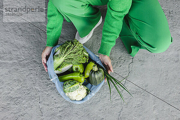Woman with mesh bag of fresh green vegetables crouching on footpath