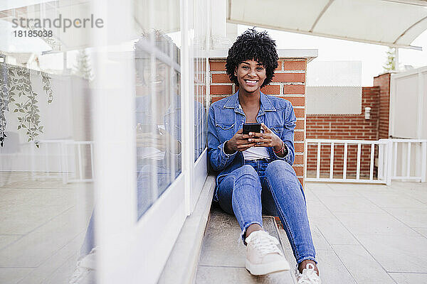 Glückliche junge Frau mit Smartphone sitzt auf der Veranda