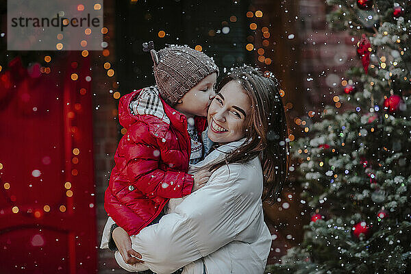Junge küsst Mutter  die vor dem Weihnachtsbaum im Schnee steht