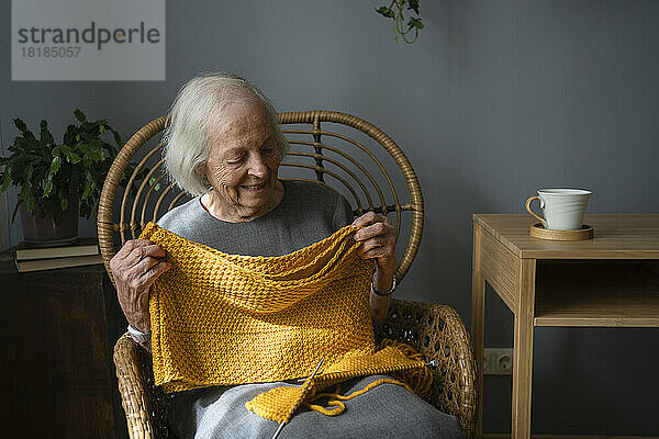 Glückliche ältere Frau hält gestrickten Schal auf Stuhl zu Hause