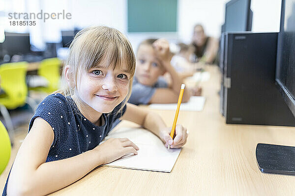 Smiling girl with pencil and book sitting at desk in classroom