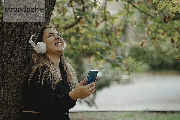 Glückliche Frau  die im Park Musik über kabellose Kopfhörer hört und ihr Smartphone in der Hand hält