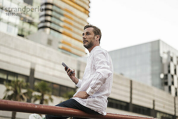 Thoughtful man sitting with smart phone on railing