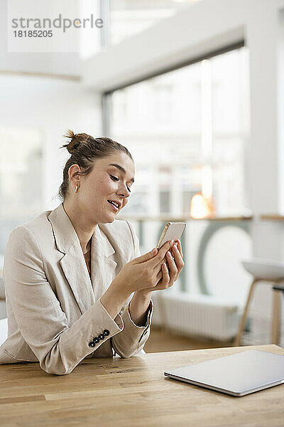 Geschäftsfrau blickt auf Mobiltelefon und sitzt mit Laptop am Schreibtisch