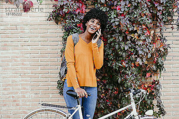 Glückliche Frau mit Fahrrad  die vor einer Efeuwand mit dem Smartphone spricht