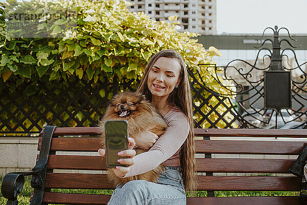 Glückliche Frau mit Hund macht Selfie per Smartphone auf Bank im Park