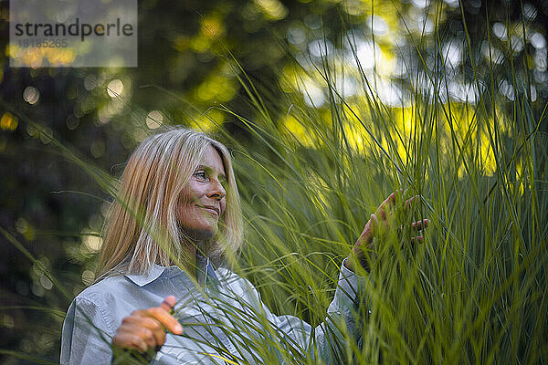 Reife Frau mit blonden Haaren berührt Pflanze im Garten