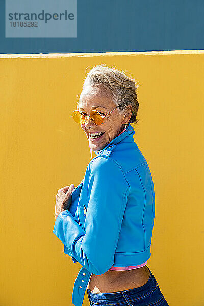 Glückliche reife Frau mit Sonnenbrille vor farbiger Wand