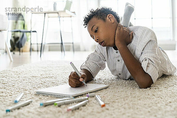 Junge schreibt mit Filzstift in ein Buch und liegt zu Hause auf dem Teppich
