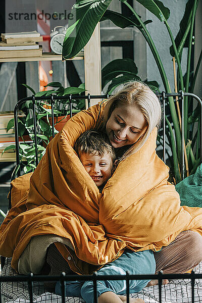 Glückliche Mutter mit Sohn  eingewickelt in eine orangefarbene Decke  zu Hause im Bett