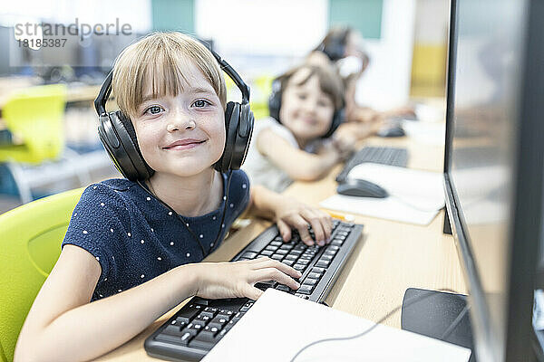 Smiling girl with bangs wearing headphones sitting at desk by computer in class