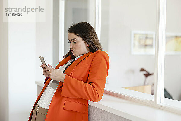 Geschäftsfrau benutzt Smartphone und stützt sich auf Schrank im Büro