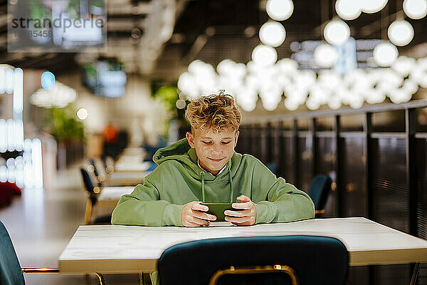 Lächelnder blonder Junge benutzt Smartphone im beleuchteten Food-Court