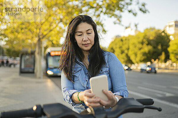 Reife Frau mietet per Smartphone auf dem Fußweg ein Fahrrad