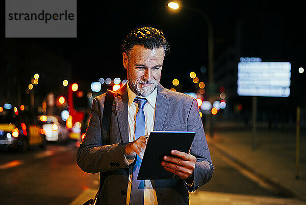 Lächelnder reifer Geschäftsmann  der nachts einen Tablet-PC nutzt