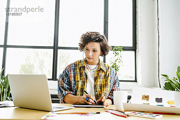 Junger Grafikdesigner sitzt mit Laptop vor dem Fenster am Schreibtisch