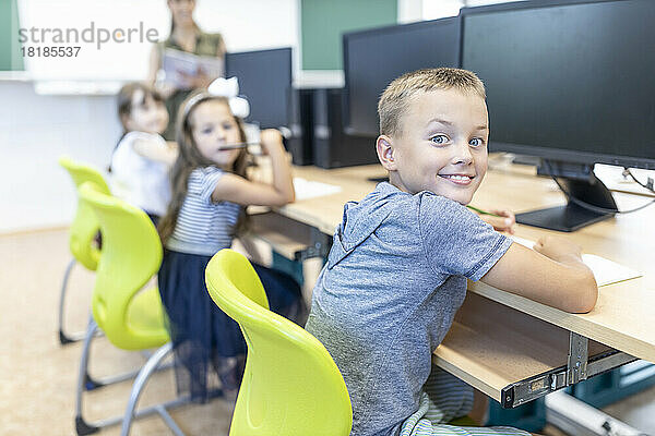 Lächelnder Junge  der im Klassenzimmer am Schreibtisch sitzt und über die Schulter schaut