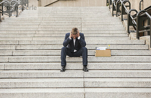 Besorgter Geschäftsmann sitzt mit Karton auf der Treppe
