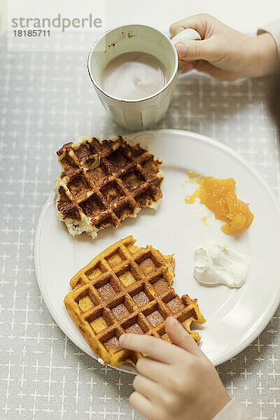 Hands of boy holding hot chocolate cup by waffles in plate on dining table