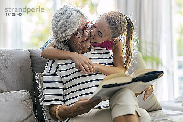 Mädchen küsst Großmutter und liest ein Buch  das zu Hause auf dem Sofa sitzt