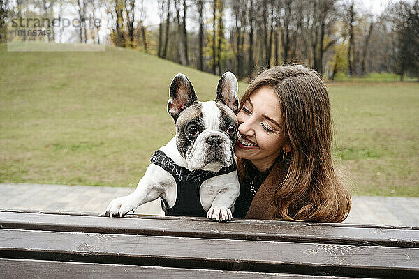 Fröhliche Frau umarmt französische Bulldogge auf Bank im Park