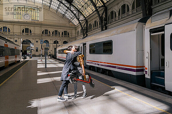 Junge Frau hält Tretroller und umarmt Freund am Bahnhof