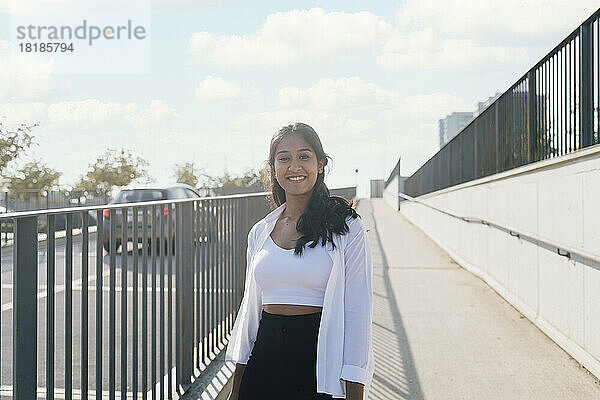 Lächelnde junge Frau steht an einem sonnigen Tag am Fußweg am Geländer