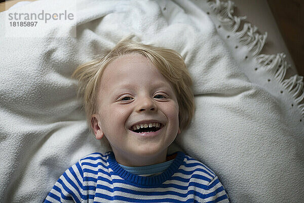 Porträt eines lachenden kleinen Jungen  der zu Hause auf einer Decke liegt