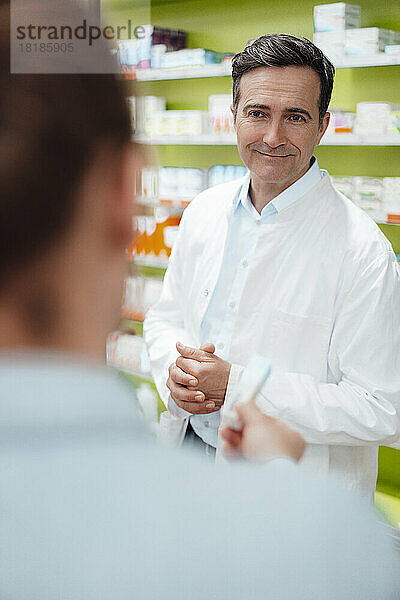 Lächelnder Apotheker blickt dem Kunden zu  der ihm Medikamente im Geschäft zeigt