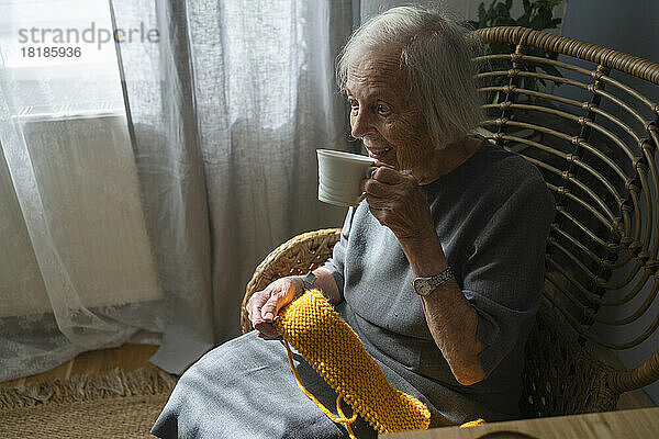 Ältere Frau trinkt Tee und hält zu Hause Strickwolle auf einem Stuhl