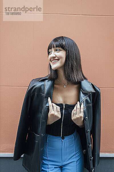 Happy young woman wearing leather jacket in front of wall