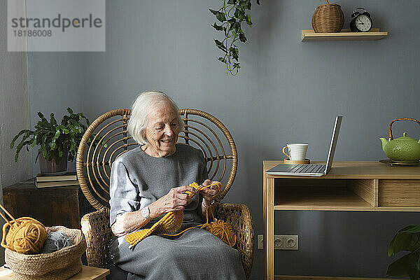 Lächelnde ältere Frau  die zu Hause auf einem Stuhl im Wohnzimmer strickt