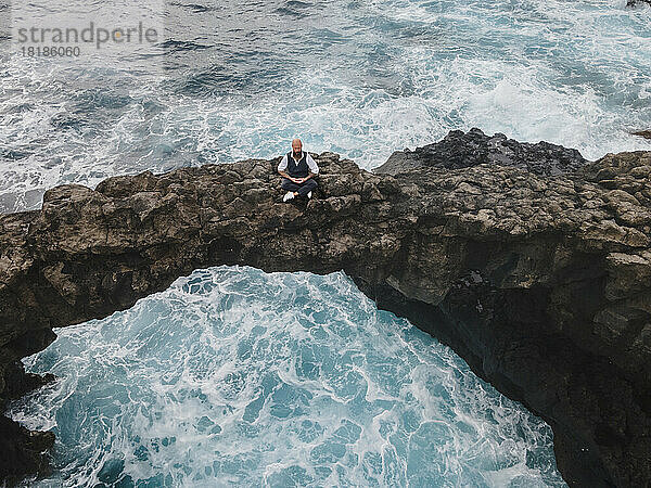 Reifer Mann praktiziert Meditation auf einem Felsbogen über dem Meer