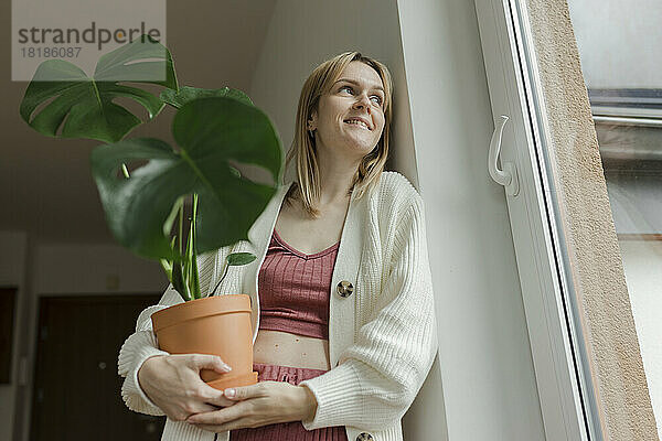 Nachdenkliche Frau hält eine Monstera-Pflanze in der Hand und lehnt zu Hause an der Wand