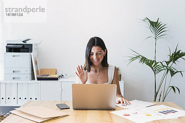Lächelnde junge Geschäftsfrau winkt per Videoanruf dem Laptop im Büro zu