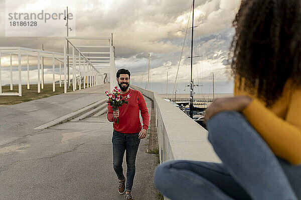 Young man walking with bouquet towards woman sitting on wall