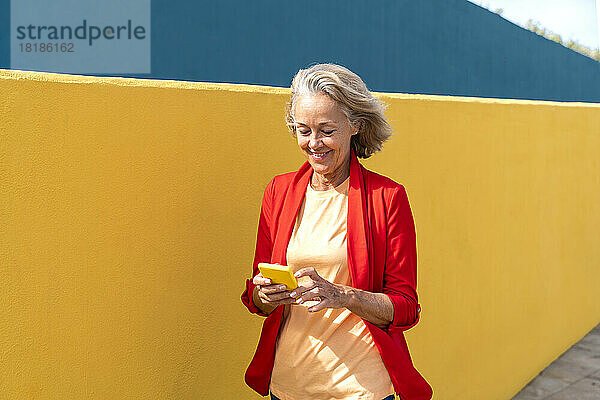 Lächelnde reife Frau  die ihr Mobiltelefon benutzt und an der gelben Wand vorbeigeht