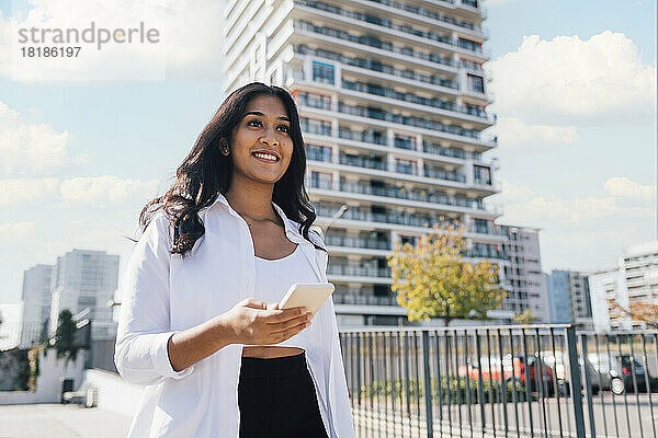 Lächelnde junge Frau mit Smartphone steht vor dem Gebäude