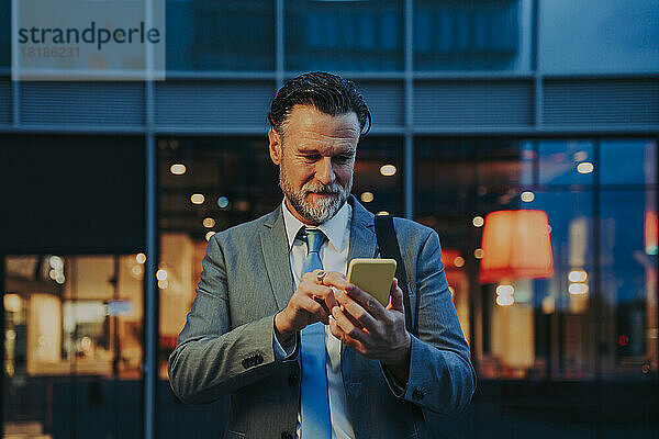 Reifer Geschäftsmann benutzt nachts Smartphone vor Gebäude